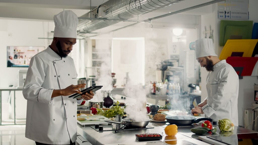 Chef using a tablet in a busy restaurant kitchen while staff prepare food during service