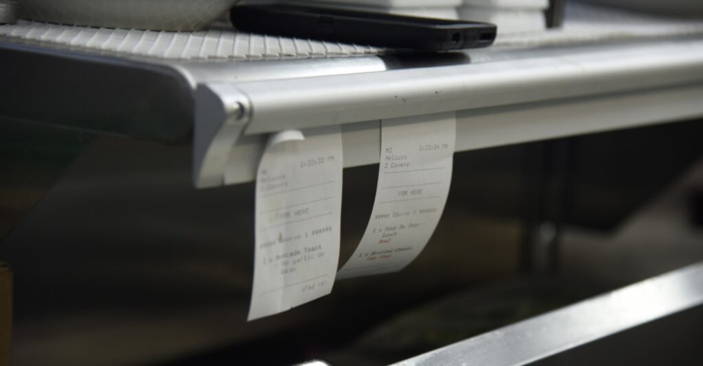 Paper tickets hanging in a restaurant back-of-house area, showing manual foodservice workflows that can become harder to manage under operational pressure.